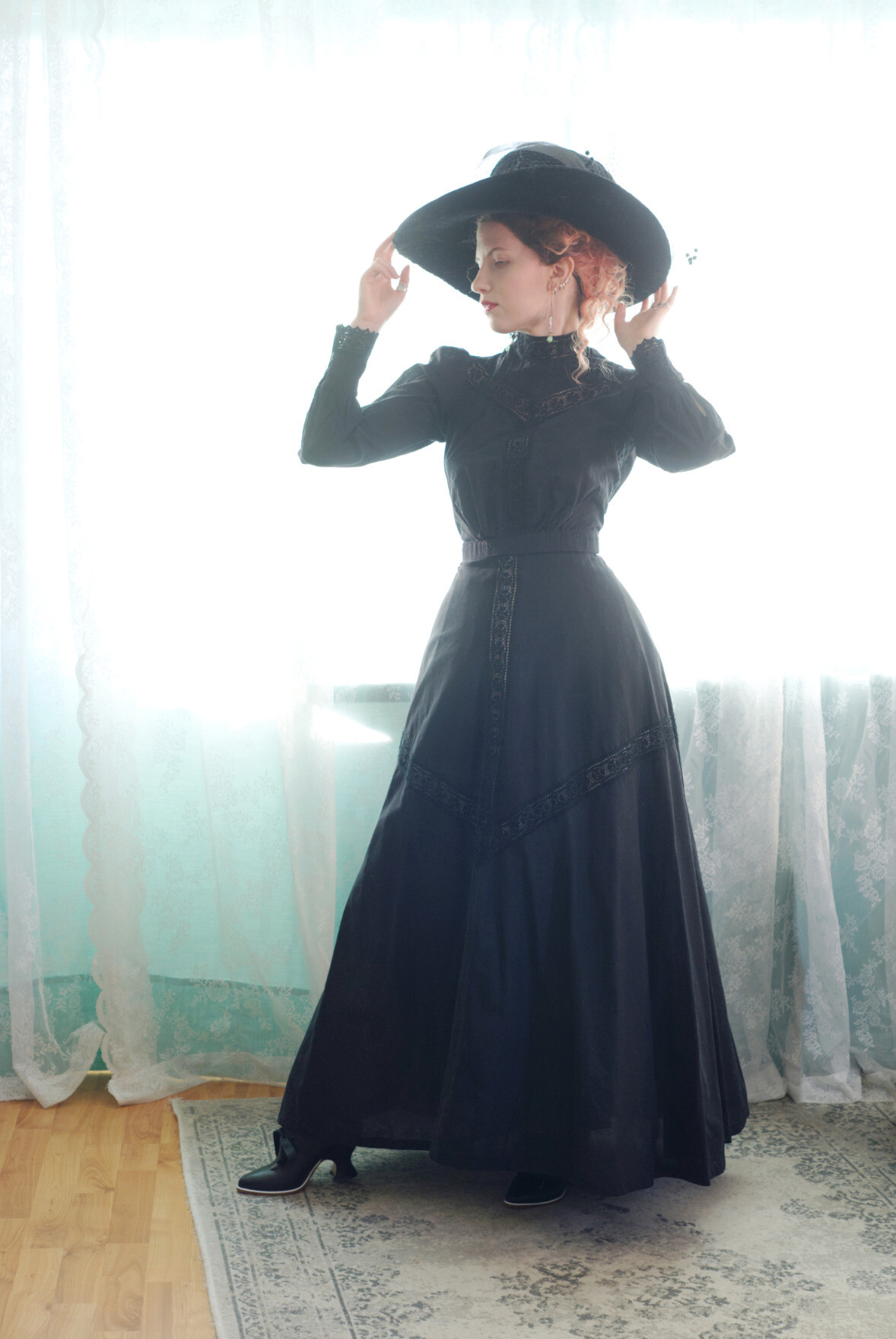A photo of Sini wearing a floor length black skirt that hugs her hips and has lace insertion details at the front and running from the centre front to centre back, rising towards the thigh and dipping down at front and back. Her hat is very large and black velvet, with dotted veiling. She is touching the brim of her hat and vibing.