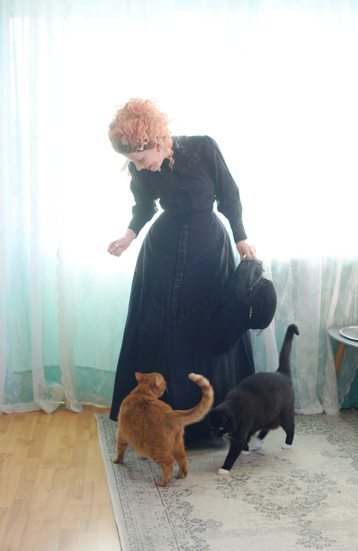 Sini smiling down and leaning down to look at cats that are sauntering in front of her. One ginger, one a black tuxedo cat. She is holding a large black hat in her hand, dressed in a black blouse and skirt with lace details. Her hips look delightfully wide in this one.