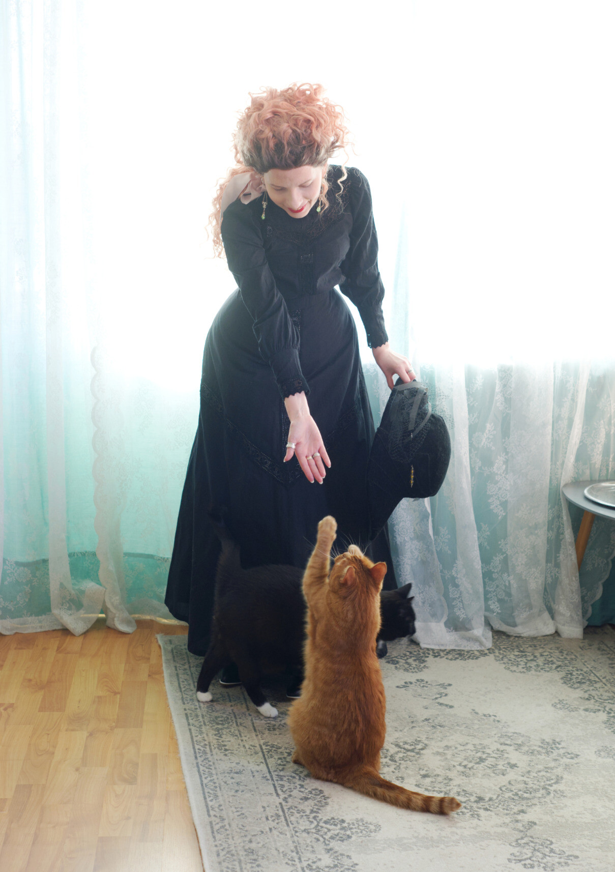 Sini, dressed in the same, stooping down a little bit to offer her open palm to a ginger cat that is standing on his back legs, his front paws reaching towards her hand, as if he is about to give a double high five. His whiskers are jutting straight forward, concentrated on Sini. This is how Karvinen shakes hands.
There is a black tuxedo cat walking just in front of the ginger one, mostly camouflaged against the black skirt, with his little white socks visible.