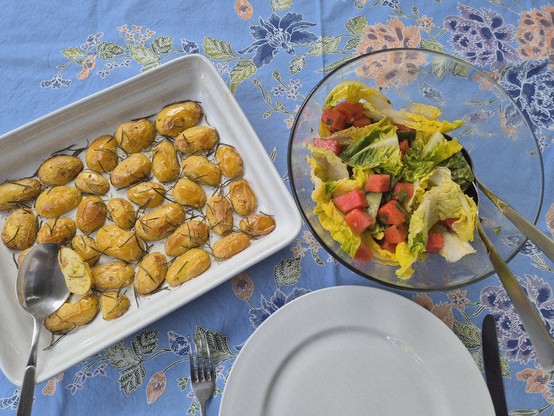 Eine Glasschüssel mit Salat, eine Auflaufform mit Kartoffelhälften von kleinen Kartoffeln mit der Schale, davor liegen ein Teller und Besteck, alles auf einer bunten Tischdecke. Es sieht sommerlich aus. 