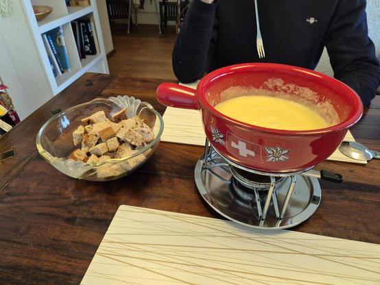 Auf einem dunklen Holztisch steht eine Schale mit Brotwürfeln, daneben auf einem Rechaud ein Fonduetopf aus Keramik, in dem geschmolzener  Käse ist. Dahinter ist der Oberkörper einer Person zu erkennen, die eine Fonduegabel in der Hand hält. 