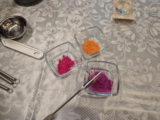 Three little glass jars with pigments (micas) in them in orange, dark pink and purple. A little Spatula rests on one of the jars. The table underneath is covered with a plastic table cloth for protection.