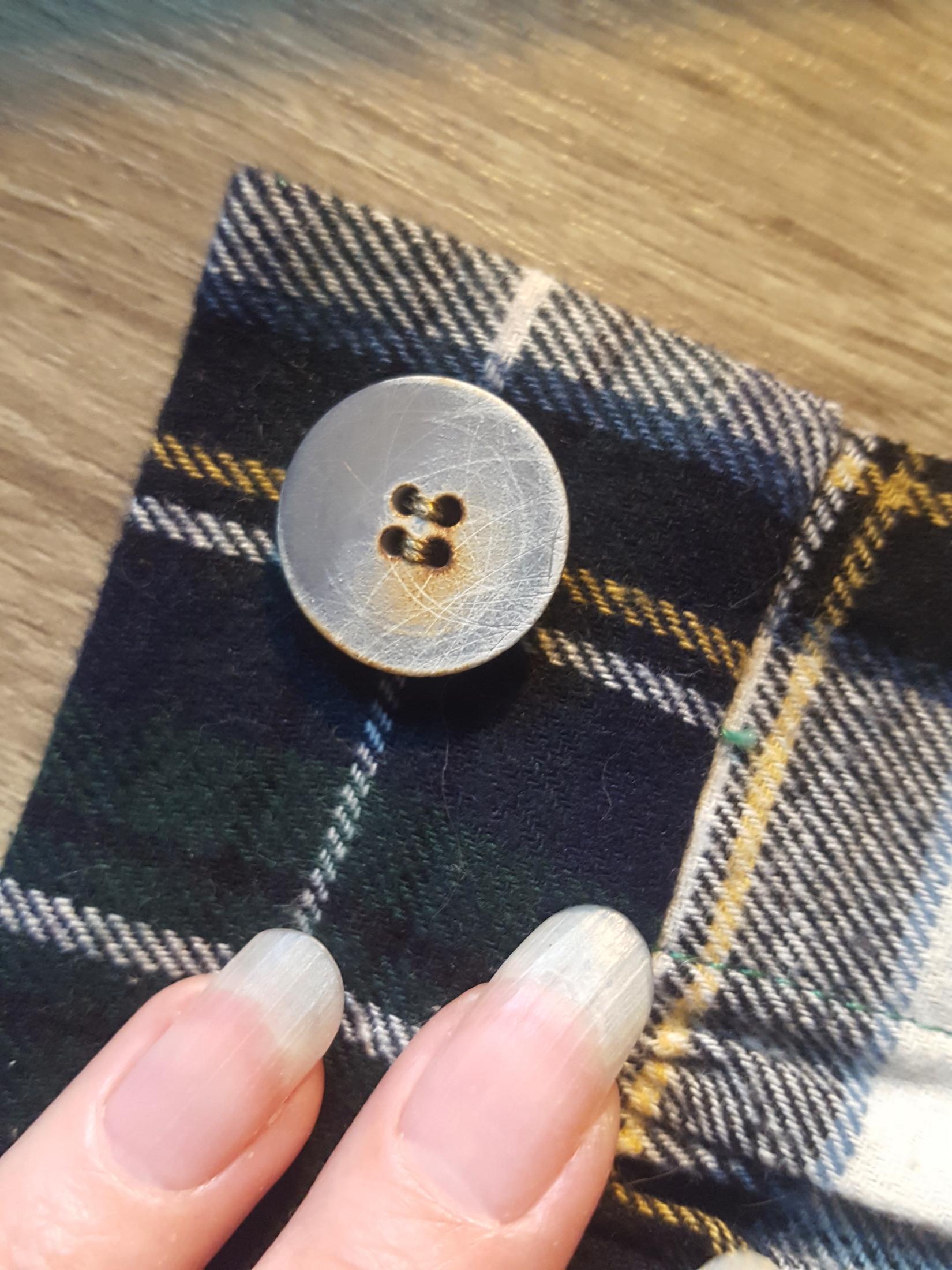 A very close up photo of the back placket of a flannel undergarment. The plaid pattern of dark green, very dark blue, lighter dark blue, a sort of gray blue and yellow goes nearly perfectly with the one singular multicoloured button sewn onto the waistband. The button has hues of the same storm grey blue, a bluish gray base, a bit of the same warm yellow and even the sort of white as a cool texture on top. It's like the button was made for the fabric, despite almost a decade separating their purchase dates. Sini's fingertips with long oval natural nails in frame for colour calibration.