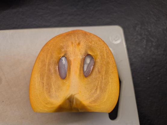 Eine halbe Kaki aufgeschnitten auf einem Schneidbrett. Die beiden Kerne sehen aus wie Augen, die besorgt blicken.