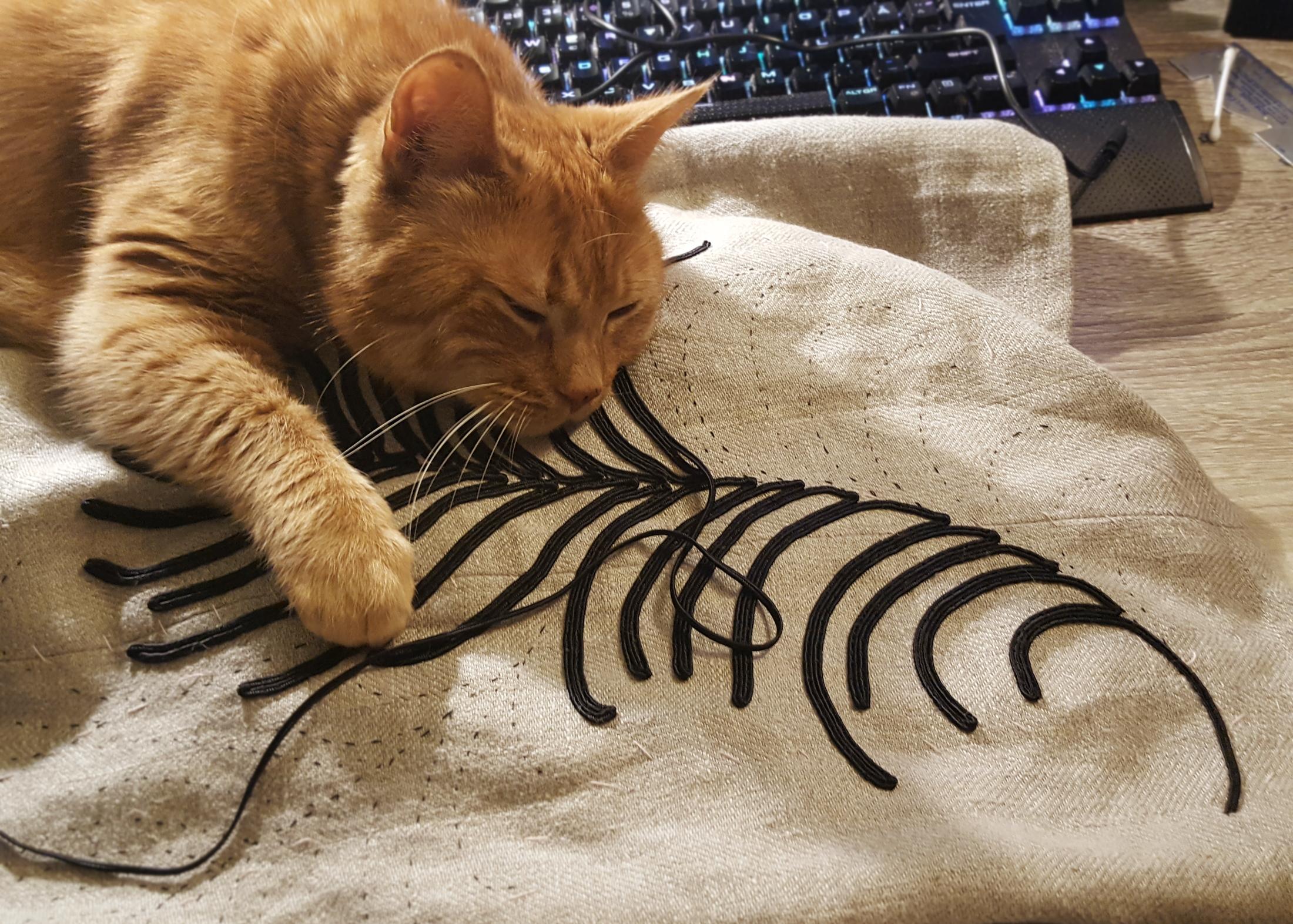 A ginger tabby is conked the heck out on a linen skirt that is getting soutache hand sewn onto it. He is determinedly napping on the very elaborate decoration, gently grasping on with claws on the raised texture of the braid. A keyboard in the background.