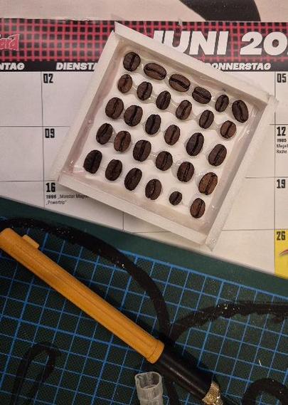 Dreißig Kaffeebohnen sind mit Heißkleber in einer kleinen Schachtel aus Schaumpappe befestigt. Die Bohnenseite mit der Rille ist jeweils oben.
Im Hintergrund eine Badtelmatte und ein Cutter.