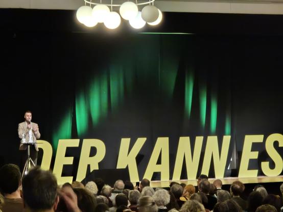Grün angestrahlter Vorhang im Hintergrund einer Bühne, davor in großen, gelben Buchstaben: "Der kann es."
Links auf der Bühne an einem Mikrofon ein Mann in hell Sacko, weißem T-Shirt und dunklen Hosen. Es ist Benjamin Bauer, Kandidat für Karlsruhe West.