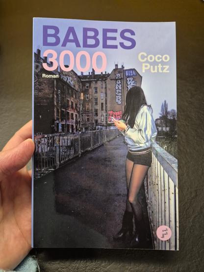 Cover eines schmalen Buches. Titel: Babes 3000, von Coco Putz, Roman. Frohmann Verlag. Titelbild: Foto einer jungen Frau, die das Gesicht von der Kamera abgewendet hat. Sie hält ein Handy und einen To-go-Becher in der Hand und lehnt an einem Brückengeländer in einer abgerockten, urbanen Umgebung. Sie trägt einen silbernen Blouson und Hotpants mit durchsichtigen Strumpfhosen und Stiefel bis zur Wade. Sie hat lange, schwarze Haare. 