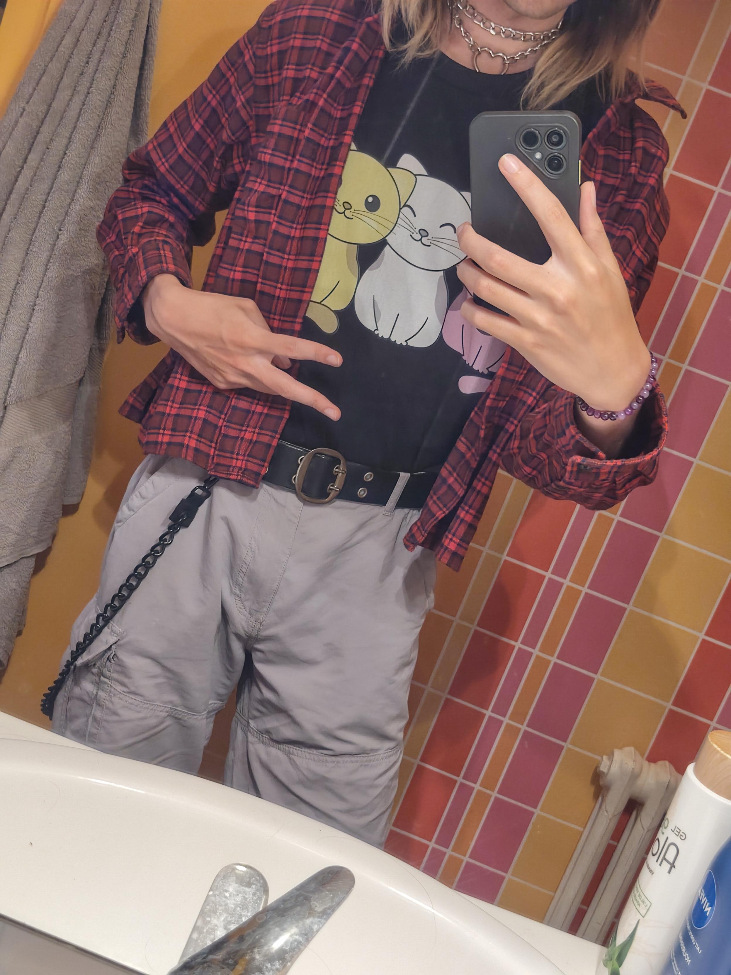Selfie of Pouet·te in its bathroom.
It wears a grey short with a large black belt and a chain attached to it, a red checkered shirt, a black T-shirt with three colored cats (yellow, white, purple) creating the enby flag, and a double chained chocker with a heart.
One of its hand form a V shape