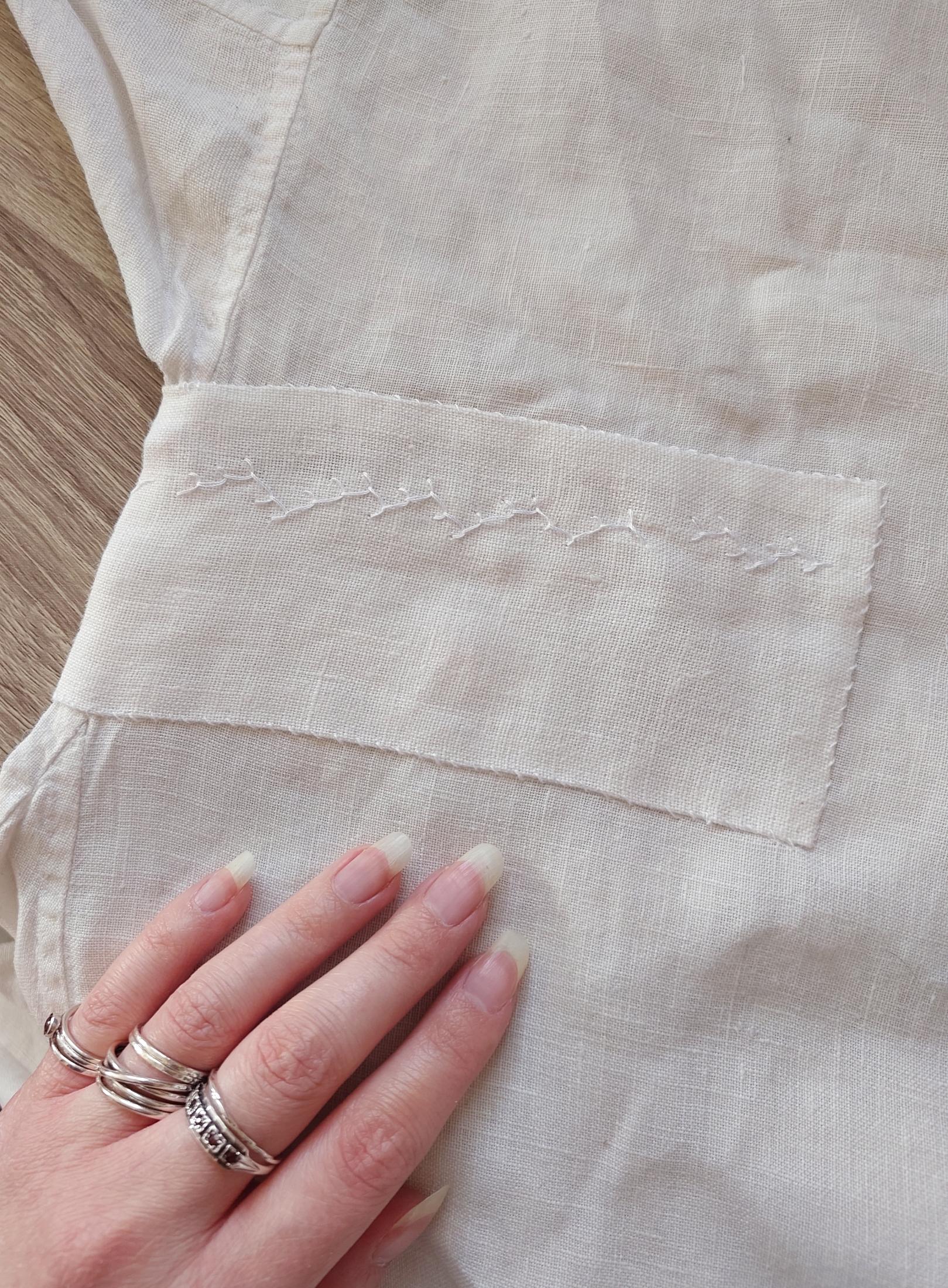 The patch, finished, sewn on with small but not tiny whipstitch in white linen thread. There's one single horizontal line of pretty erratic feather stitch going along the patch, right where the torn section is, to keep the bits under the patch from flapping about. Sini's pale hand in frame again, ooh the silver rings are so shiny, makes the patch look a lot more intentional and fine than it is!