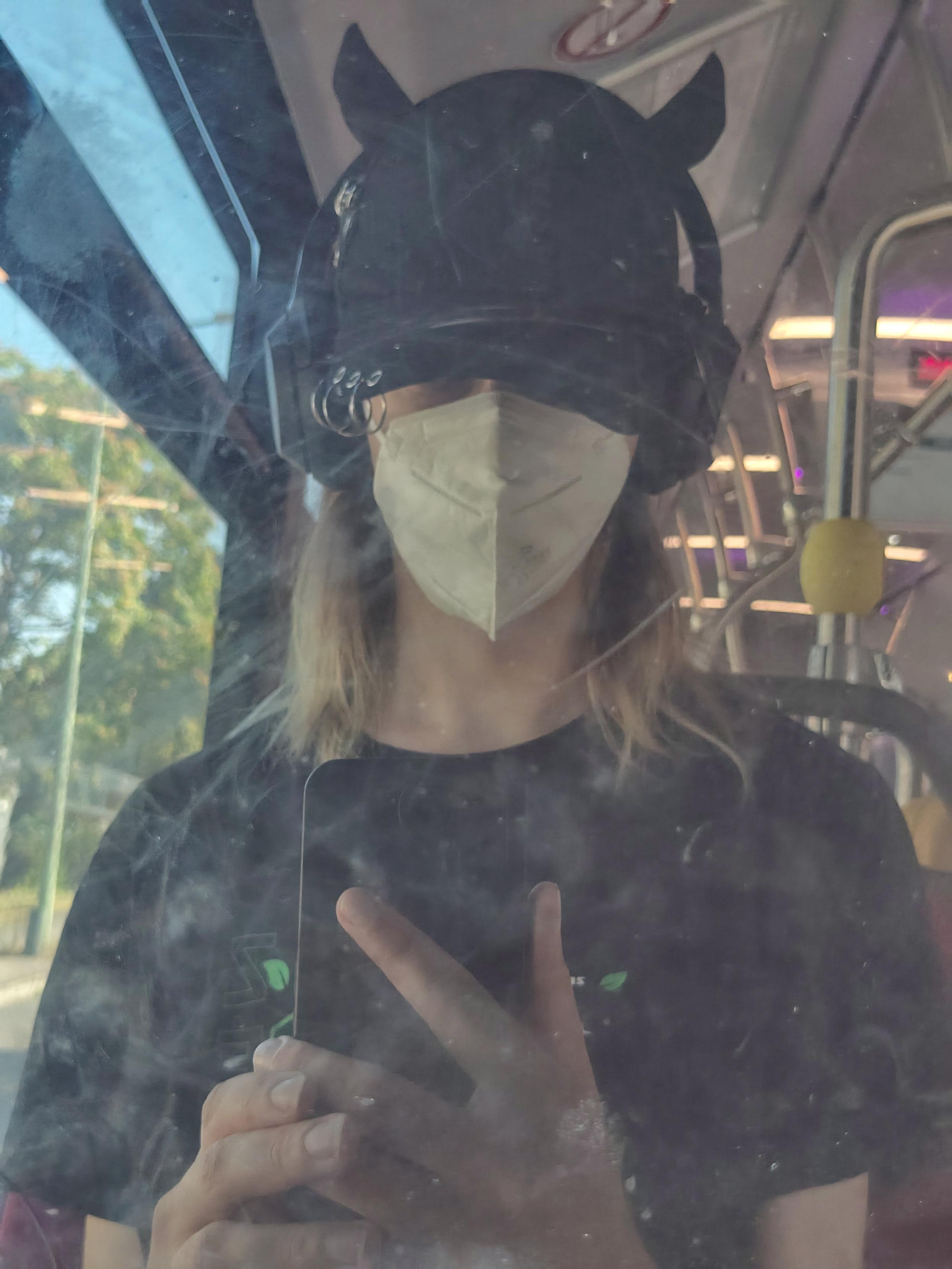 Poor quality selfie of Pouet·te through a bus's window
It wears a black shirt, a white FFP2 mask, a black headphone and a black cap with two little horns
Its eyes are covered by the cap