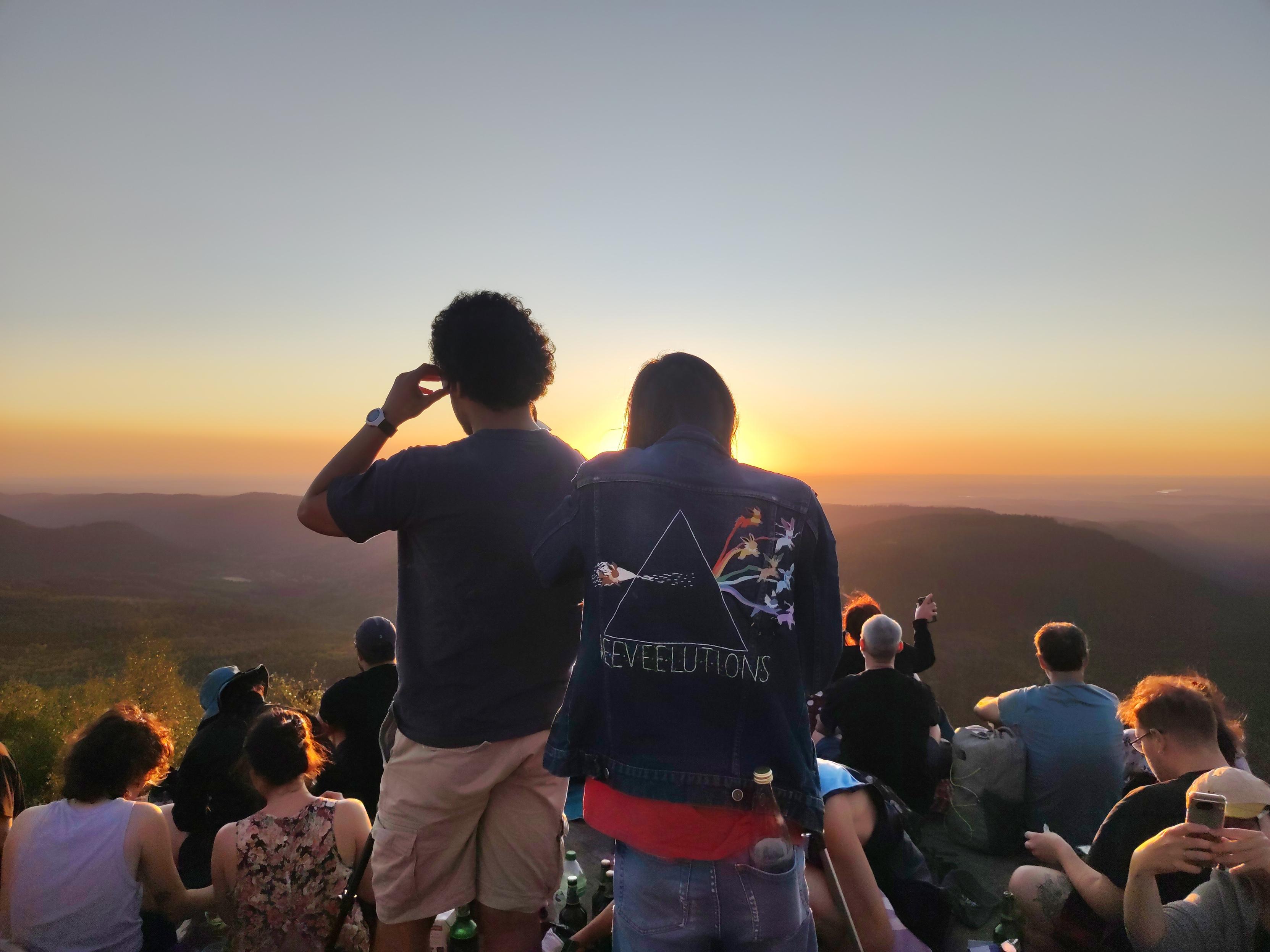 des gens qui regardent ou prennent en photo l'éclipse dont 1 à contre-jour porte une veste en jean brodée d'un prisme au travers passe le pokemon Evoli pour évoluer en toutes ses formes possibles