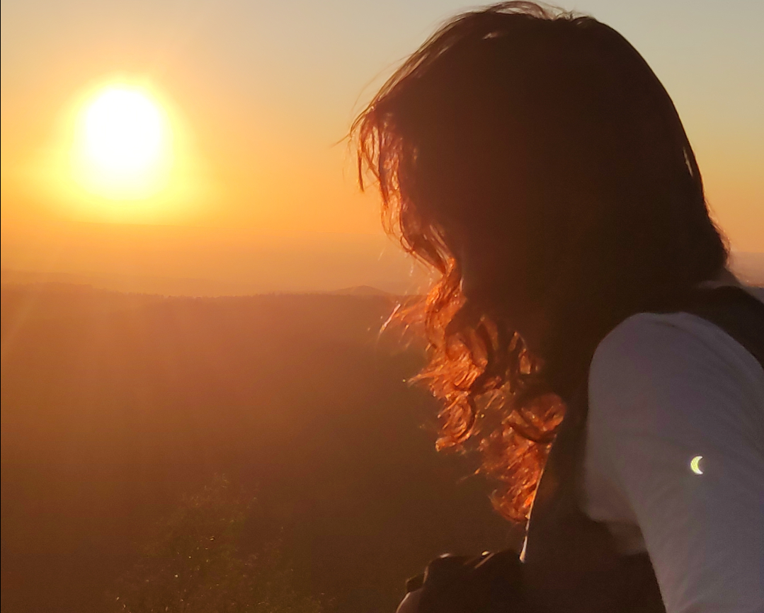 une personne de profil regardant l'éclipse au coucher du soleil, colorant ses cheveux d'une lumière orangée. sur son bras, un reflet de lentille provoque l'apparition de l'éclipse très nette en surimpression