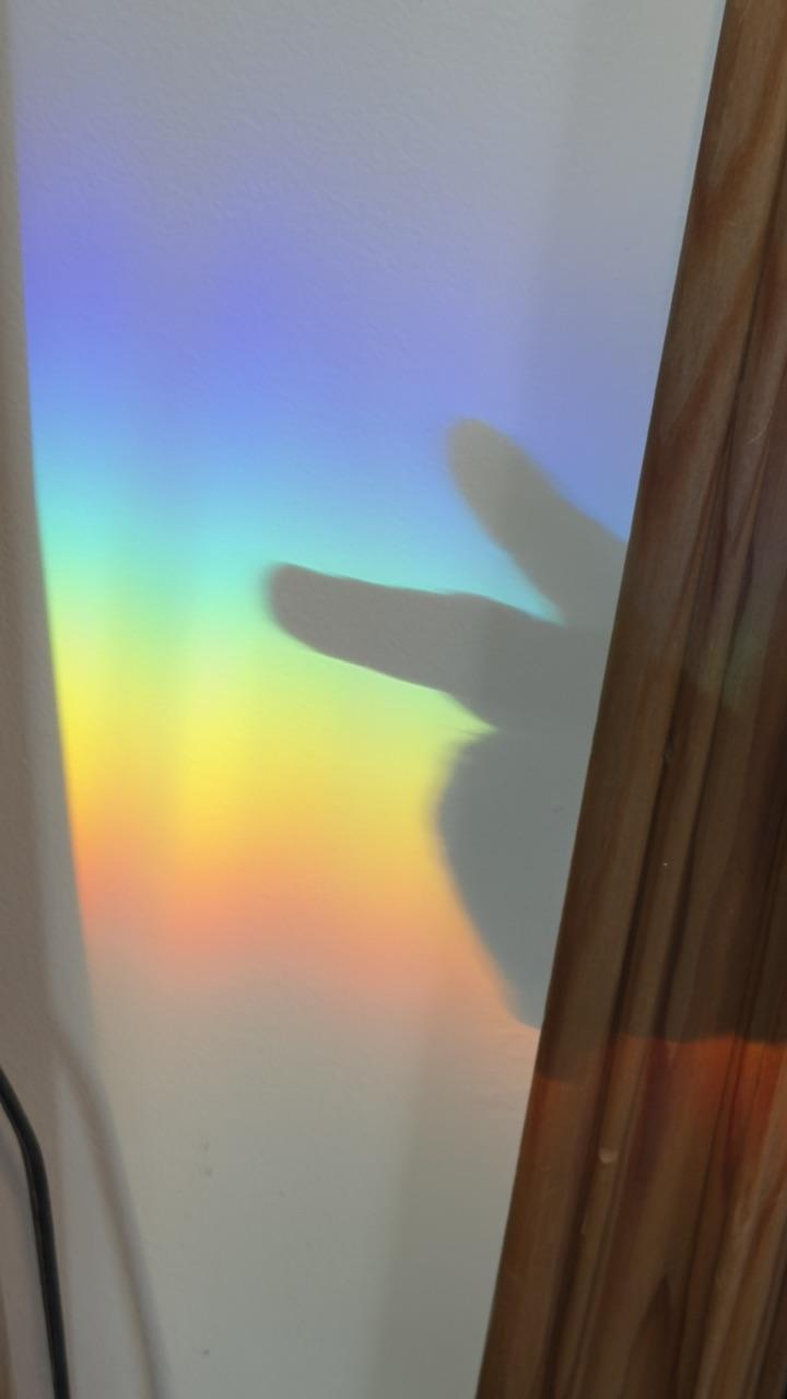 Light passing through a piece of glass in a window has generated a rainbow on a white wall. Somebody is making a peace symbol with their hand that looks vaguely like a rabbit shadow.