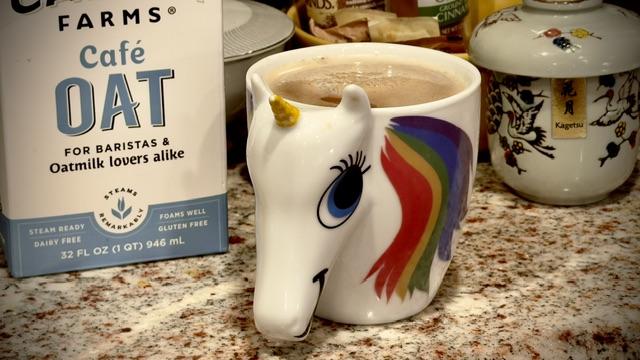 Shows a unicorn shaped coffee mug with a rainbow main and blue eyes, sporting a smile and a yellow horn. Inside is whitened coffee. It is on a brown speckled whitegranite countertop. In the background you can see a Japanese mug and a box of oat milk.