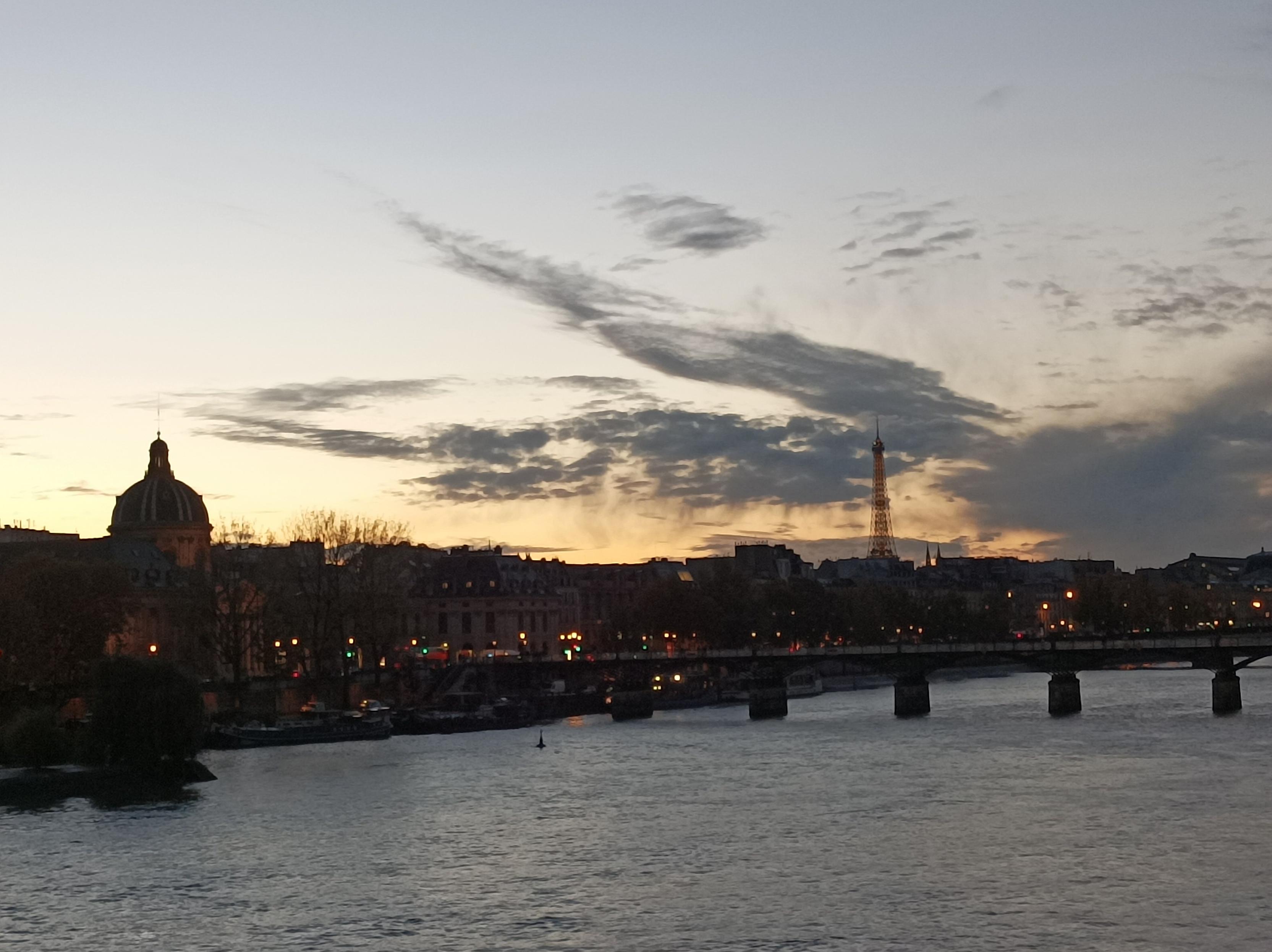 Photo au coucher du soleil depuis un pont parisien de la Seine, au loin le pont des arts et la tour eiffel, ciel clair, avec quelques nuages au loin