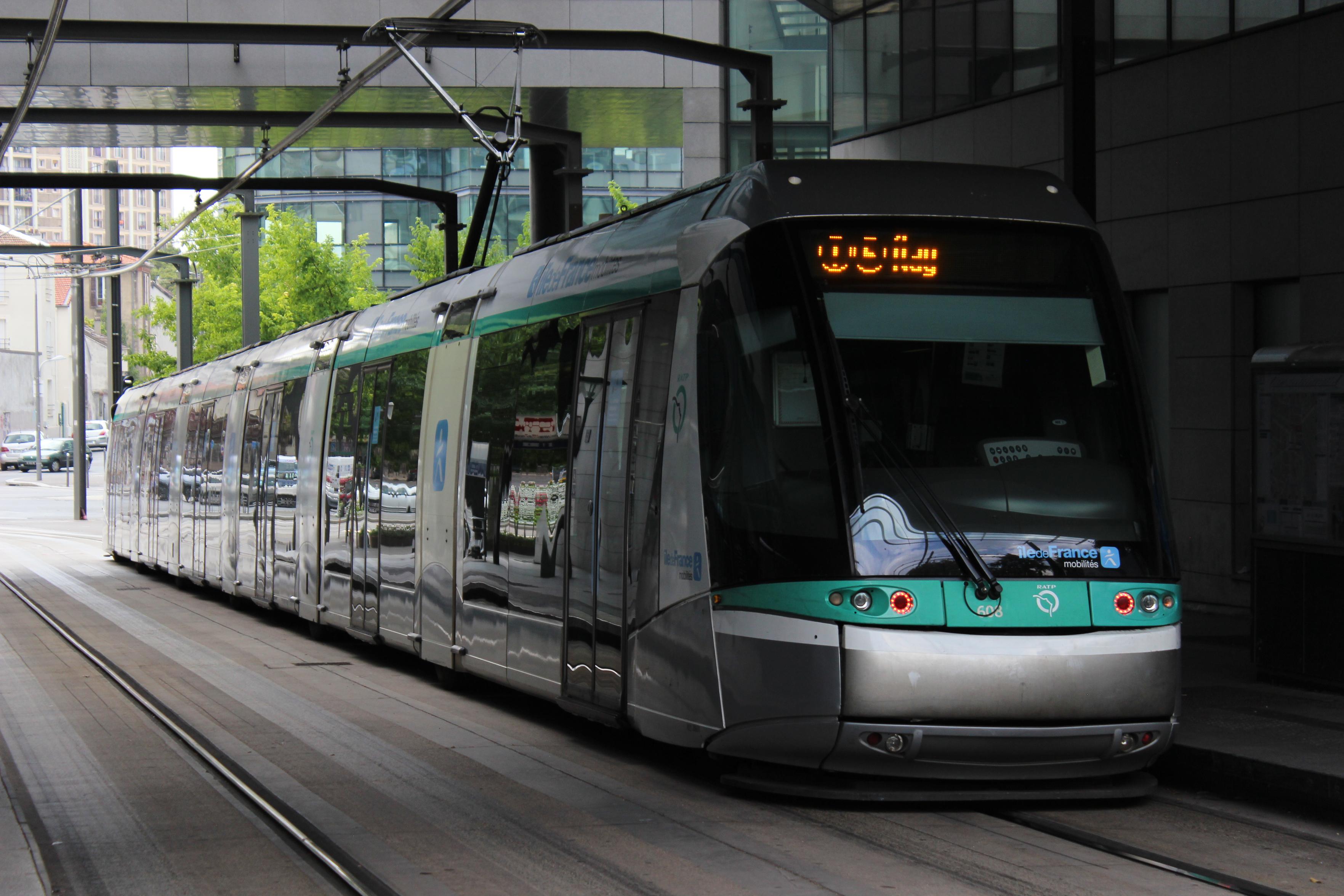 Photo plutôt sombre d'un tramway gris, blanc et bleu vert prise légèrement de biais. Il est partiellement sous une structure mais au bout on voit une rue non couverte (il semble faire gris).
