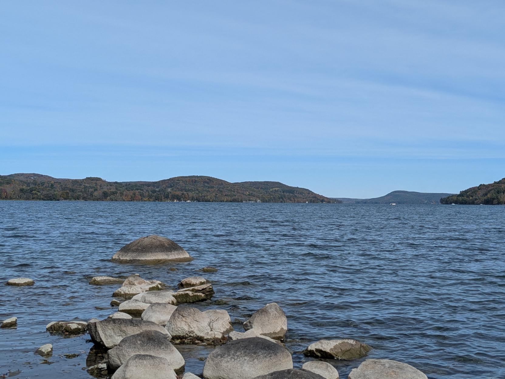 Wide angle view of the lake. Clouds streak the blue sky and the low rounded hills are mottled green, yellow, brown, orange. A line of rocks extends from the foreground shore into water that's slightly choppy.